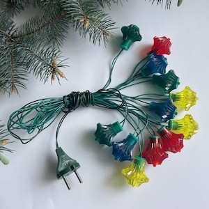 May include: A string of colourful Christmas lights with red, blue, and yellow star-shaped bulbs. The lights are connected by green wires and have a green plug. The lights are arranged on a white surface with a sprig of evergreen.