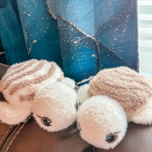 May include: Two plush turtle toys crafted from soft, fuzzy yarn. Each turtle features a white head and flippers paired with a tan and white striped shell. The creatures have dark, glossy eyes and a rounded, stuffed body design with a textured finish.