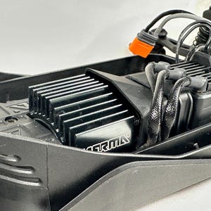 May include: Close-up of a black electric motor with a silver heatsink and the word "GRIMA" printed on it. The motor is mounted in a black plastic chassis with wires and connectors.