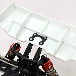 Puede incluir: Un alerón blanco de varias secciones con un soporte de montaje negro está fijado a un chasis negro. Los componentes de suspensión rojos y negros son visibles debajo del alerón. Este es un componente para un coche radiocontrolado.