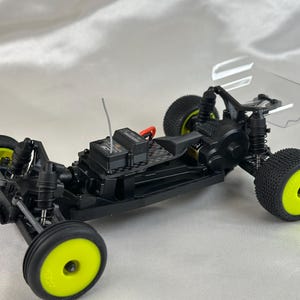 以下が含まれることがあります： 透明なプラスチック製のボディを持つ、黒と黄色のラジコンカーのシャーシ。シャーシにはカーボンファイバープレートと黒いモーターが搭載されています。車は、黒いタイヤを履いた黄色のホイールを備えています。