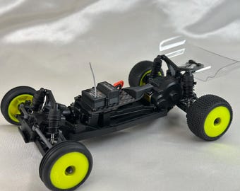 May include: A black and yellow radio-controlled car chassis with a clear plastic body. The chassis has a carbon fibre plate and a black motor. The car has yellow wheels with black tyres.