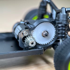 May include: Close-up of a black and silver electric motor with a white gear. The motor is attached to a black chassis with a black tire and a green rim.