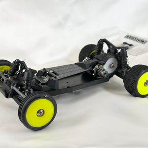 May include: A black and yellow RC car chassis with a clear plastic wing. The chassis has a carbon fiber plate and a gear differential.