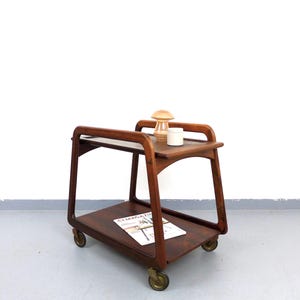Puede incluir: Carro de bar de madera oscura de estilo moderno de mediados de siglo, con dos estantes y ruedas de latón. Una pequeña lámpara y una taza blanca están en el estante superior. Una revista está en el estante inferior.