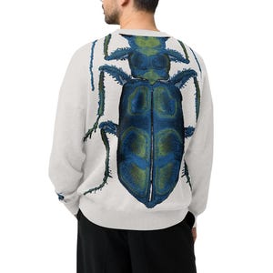 May include: A light gray long-sleeve crewneck sweater featuring a large, detailed graphic print of a blue and green beetle on the back. The sweater is paired with loose-fitting black trousers and black shoes, shown from a rear-facing perspective.