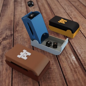 Puede incluir: Una caja de dados azul con una torre de dados negra y dorada. La caja está abierta y tiene dos dados negros dentro, uno con un 5 y el otro con un 4. La torre de dados está cerrada y tiene un logotipo de castillo negro y dorado en la parte superior.