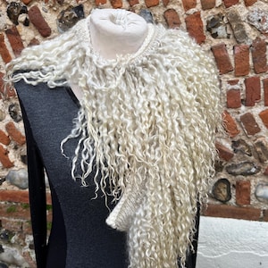 May include: A cream-colored, curly wool scarf displayed on a mannequin. The scarf has a textured, shaggy appearance with long, flowing strands. The mannequin is wearing a dark gray top, and the background is a brick wall.