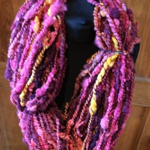 Könnte beinhalten: Ein farbenfroher, handgestrickter Schal in den Farben Rosa, Lila und Gelb. Der Schal ist aus verschiedenen Garnen gefertigt und hat eine einzigartige, strukturierte Optik.