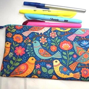 May include: A rectangular fabric pencil case featuring a dark blue background with a colorful pattern of birds and flowers in shades of orange, yellow, pink, and light blue, accompanied by four pens and markers in blue, yellow, and pink.