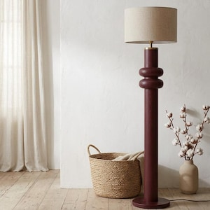 May include: A floor lamp with a deep burgundy wooden base featuring two rounded, stacked decorative rings on the central column. The lamp is topped with a cylindrical, light beige fabric lampshade and has a circular base resting on the floor.