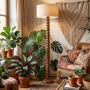 May include: A floor lamp with a tall, stacked wooden sphere base and a cylindrical beige fabric shade. The lamp stands upright, emitting a warm glow, positioned among several potted green plants and a rattan chair with patterned cushions.