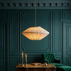 May include: A pendant light fixture featuring a wide, pleated, off-white fabric shade with a brass-toned metal frame and a matching thin vertical rod that connects to a circular ceiling mount, emitting a warm, soft glow from within the shade.