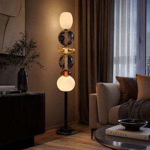 May include: A modern floor lamp with a black metal pole and a marble base. The lamp features a series of glass orbs and rings in white, clear, amber, and smoky gray. The lamp is illuminated, casting a warm glow in the room.