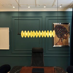 May include: A modern office space featuring a black marble desk, a black leather chair, and a unique light fixture. The light fixture is a series of oval-shaped, warm-toned lights suspended from the ceiling. A painting of a woman in a head wrap hangs on the wall.