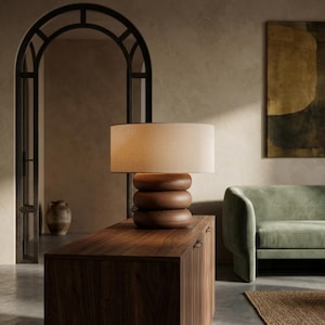 May include: A table lamp featuring a cylindrical, off-white fabric shade resting on a base composed of three stacked, rounded, dark brown wooden rings. The lamp emits a soft, warm glow, highlighting the smooth, polished texture of the wood.