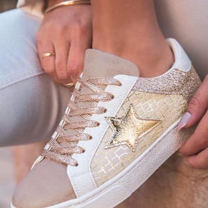 Op de afbeelding: Crèmekleurige sneakers met een gouden sterapplicatie. De schoenen hebben een beige suède neus, witte leren accenten en gouden glitter op de hiel. De veters zijn goudkleurig.