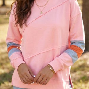 May include: A long-sleeve crewneck sweatshirt in light pink featuring a diagonal seam across the chest and color-blocked sleeves with horizontal stripes in blue, orange, and light pink. The sweatshirt has ribbed cuffs and a straight hem with side slits.