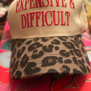 Puede incluir: Gorra de béisbol beige con las palabras "EXPENSIVE & DIFFICULT" bordadas en rojo. La visera presenta un estampado de leopardo. Este sombrero es un accesorio elegante.