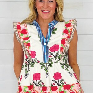 May include: A sleeveless top with a floral print. The top features a white base with a pattern of pink flowers and green leaves. The neckline and armholes are trimmed with striped fabric. The top has a blue and white striped collar.