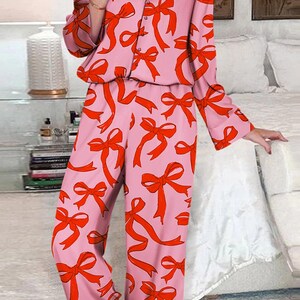 May include: Pink pajama set with a red bow pattern. The long-sleeved top has a button-down front and the pants are full-length. The sleepwear is made of a soft material and is suitable for lounging or sleeping.