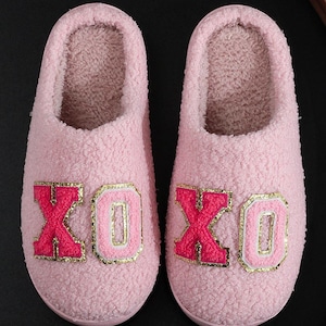 May include: A pair of pink plush slippers with the letters "XO XO" embroidered in red and gold. The slippers are designed for indoor wear and have a soft, comfortable appearance.