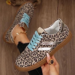 May include: Leopard print sneakers with light blue laces and a brown sole. The shoes have a white interior and metallic silver accents. The sneakers are a stylish and trendy footwear option.