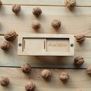 May include: A wooden box with a slot for holding nuts. The box has the word "Available" engraved on the front. The box is surrounded by walnuts on a wooden surface.