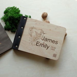 May include: A wooden photo album with a black leather spine and silver rivets. The cover is engraved with a hummingbird and floral design, along with the names "James & Emily" and the date "25.05.2024".