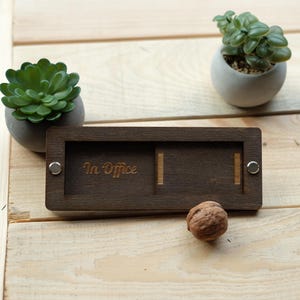 Puede incluir: Un letrero de madera marrón oscuro con un panel deslizante. El letrero dice "In Office" y tiene un panel en blanco que se puede deslizar hacia la izquierda o hacia la derecha para indicar si alguien está en la oficina o no.