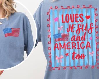 May include: A dusty blue t-shirt with a small American flag on the pocket and a larger design on the back. The back design includes an American flag, the text "LOVES JESUS and AMERICA too", stars, hearts, and bows.