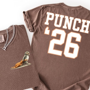 May include: Two brown t-shirts. One is folded, displaying a monkey graphic and a silver chain necklace. The other shirt has "PUNCH" and the number "26" printed on the back in white with orange accents. Both shirts are on a white background.