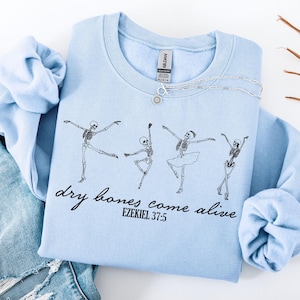 May include: A light blue sweatshirt with a graphic of dancing skeletons, the text "dry bones come alive" and Ezekiel 37:5. The sweatshirt is paired with light blue jeans and a silver necklace.