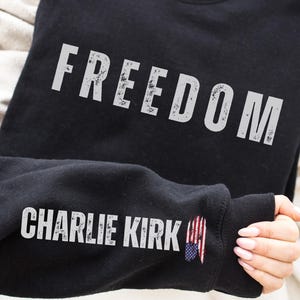 以下が含まれることがあります： 「FREEDOM」の文字がかすれた白い文字でプリントされた黒いスウェットシャツ。袖には「CHARLIE KIRK」の文字と小さなアメリカ国旗のグラフィックが施されています。スウェットシャツはニュートラルカラーの生地の上に展示されています。