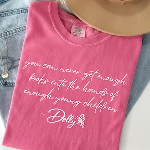 May include: A pink short-sleeved t-shirt featuring the white text: you can never get enough books into the hands of enough young children - Dolly, accompanied by a butterfly graphic. The shirt is paired with light blue denim jeans and a tan hat.