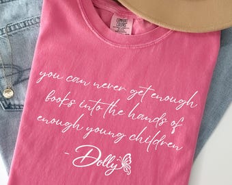 May include: A pink short-sleeved t-shirt featuring the white text: you can never get enough books into the hands of enough young children - Dolly, accompanied by a butterfly graphic. The shirt is paired with light blue denim jeans and a tan hat.