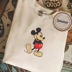 May include: A beige sweatshirt with an embroidered Mickey Mouse design. The cartoon character is depicted in his iconic outfit, with black ears, red shorts, and yellow shoes. A circular patch with the word "EMBROIDERY" is also visible.