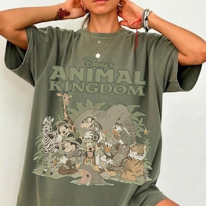 May include: A moss green t-shirt featuring the text "Disney's Animal Kingdom" above a graphic of various cartoon animals. The graphic includes a zebra, giraffe, elephant, tiger, and other characters. The shirt has short sleeves and a relaxed fit.