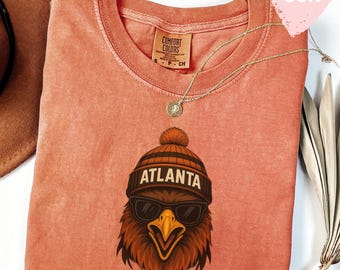 May include: A coral-coloured Comfort Colors sweatshirt with a graphic of a cool eagle wearing sunglasses and a beanie with "ATLANTA" printed on it. A gold necklace and a brown hat are also visible. The shirt has a "super soft" heart-shaped graphic.