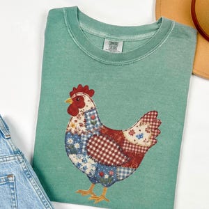 Peut inclure: Un t-shirt vert d'eau Comfort Colors avec un motif de poulet patchwork. Le poulet a une crête rouge, un bec jaune et un corps composé de tissus à motifs rouges, bleus et blancs. Un jean bleu et un chapeau beige sont également visibles.