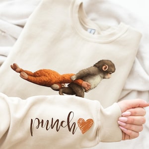 May include: Beige sweatshirt featuring a whimsical illustration of a brown monkey carrying an orange plush toy. The sleeve displays the word "punch" in a script font, accompanied by a brown heart graphic. The sweatshirt is folded on a white surface.