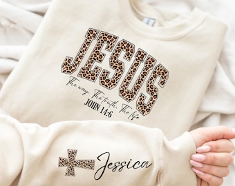 May include: Beige sweatshirt with the word "JESUS" in a leopard print design. The text "The way. The truth. The life. JOHN 14:6" is below the word. The sleeve features a leopard print cross and the name "Jessica".