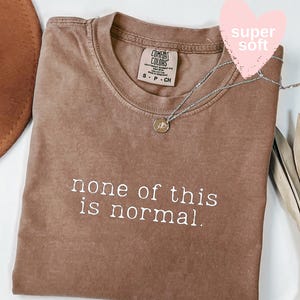 May include: Espresso-colored t-shirt with the text "none of this is normal" in white. The shirt has a crew neck and a small tag at the neckline. A silver necklace with a pendant is draped across the shirt. A pink heart-shaped sticker says "super soft".