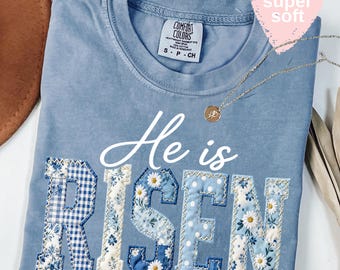 Op de afbeelding: Lichtblauwe t-shirt met de tekst "He is Risen" in witte letters en patchwork letters. De letters zijn gemaakt van blauwe en witte bloemen- en ruitpatronen. Een gouden ketting en een roze hartvormig label zijn ook zichtbaar.