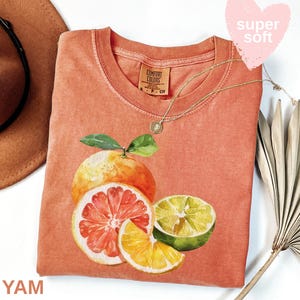 Op de afbeelding: Koraalrood Comfort Colors T-shirt met een aquarel illustratie van citrusvruchten: een grapefruit, een limoen en een sinaasappelschijf. Een gouden ketting en een bruine hoed zijn zichtbaar. De tekst "super soft" staat in een roze hart.