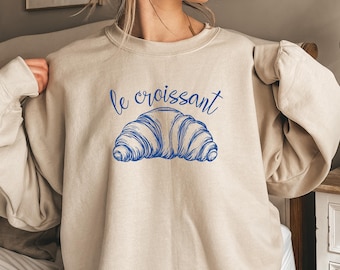 May include: Beige sweatshirt with the blue text "le croissant" above a blue illustration of a croissant. The sweatshirt is a neutral color and has long sleeves.