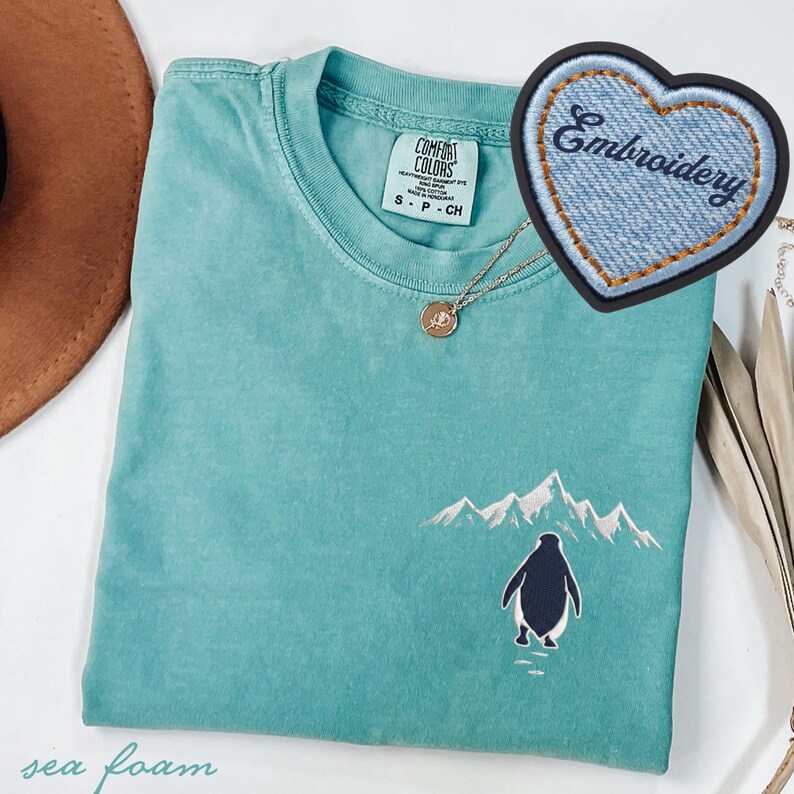 May include: Seafoam green t-shirt with an embroidered penguin and mountain scene. The shirt has a crew neck and short sleeves. A gold necklace and a heart-shaped patch that says "Embroidery" are also visible.