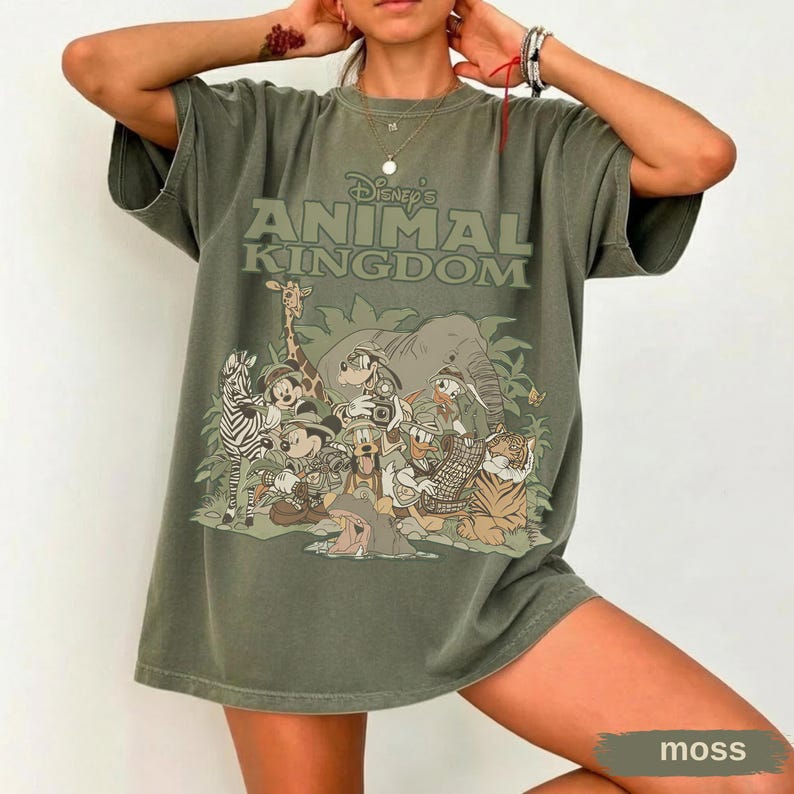 May include: A moss green t-shirt featuring the text "Disney's Animal Kingdom" above a graphic of various cartoon animals. The graphic includes a zebra, giraffe, elephant, tiger, and other characters. The shirt has short sleeves and a relaxed fit.