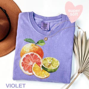 Op de afbeelding: Een violet Comfort Colors T-shirt met een aquarel illustratie van citrusvruchten, waaronder een sinaasappel, een grapefruit en een limoen. Het shirt heeft een ronde hals en korte mouwen. Een gouden ketting met een hanger is over het shirt gedrapeerd. Het woord "VIOLET" staat onderaan.