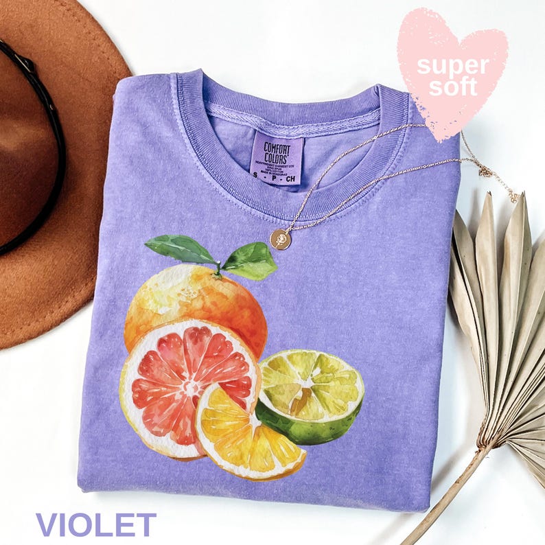 Op de afbeelding: Een violet Comfort Colors T-shirt met een aquarel illustratie van citrusvruchten, waaronder een sinaasappel, een grapefruit en een limoen. Het shirt heeft een ronde hals en korte mouwen. Een gouden ketting met een hanger is over het shirt gedrapeerd. Het woord "VIOLET" staat onderaan.